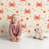 Lovely child standing in front of Whimsy Tots Crabby Coral Dance Wallpaper in Sand with crab, starfish, shell, coral, sand in design.