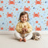 Lovely child standing in front of Whimsy Tots Crabby Coral Dance Wallpaper in Sky Blue with crab, starfish, shell, coral, sand in design.