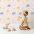 Lovely child standing in front of Whimsy Tots Sun and Cloud Kiss Wallpaper in Blush Pink with sun, cloud, raindrop, flower, star in design.