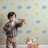 Lovely child standing in front of Whimsy Tots Sun and Cloud Kiss Wallpaper in Mint Green with sun, cloud, raindrop, flower, star in design.