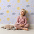 Lovely child standing in front of Whimsy Tots Sun and Cloud Kiss Wallpaper in Lavender with sun, cloud, raindrop, flower, star in design.