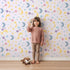 Lovely child standing in front of Whimsy Tots Starlit Wishes Wallpaper in Lavender with moon, star, constellation, shooting star, galaxy in design.
