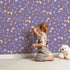 Lovely child standing in front of Whimsy Tots Starglow Constellation Dance Wallpaper in Plum with moon, star, constellation, shooting star, galaxy in design.