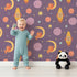 Lovely child standing in front of Whimsy Tots Fox and Koala Space Mission Wallpaper in Plum with fox, koala, moon, rocket, star, planet in design.