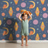 Lovely child standing in front of Whimsy Tots Fox and Koala Space Mission Wallpaper in Twilight Blue with fox, koala, rocket, moon, star, planet in design.