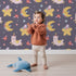 Lovely child standing in front of Whimsy Tots Llama Moon Parade Wallpaper in Twilight Plum with llama, moon, star, flower, hat in design.
