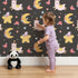 Lovely child standing in front of Whimsy Tots Llama Moon Parade Wallpaper in Midnight Black with llama, moon, star, flower, hat in design.