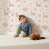 Lovely child standing in front of Whimsy Tots Celestial Petal Dreams Wallpaper in Soft Blush Pink with moon, flower, heart, star, wing in design.