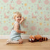 Lovely child standing in front of Whimsy Tots Celestial Petal Dreams Wallpaper in Mint Sky Green with moon, flower, heart, star, wing in design.