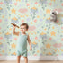 Lovely child standing in front of Whimsy Tots Starlight Blossom Dreams Wallpaper in Mint Sky Green with star, flower, cloud, leaf in design.