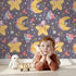 Lovely child standing in front of Whimsy Tots Starry Cuddle Sky Wallpaper in Twilight Plum with star, moon, flower, hat in design.