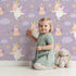 Lovely child standing in front of Whimsy Tots Unicorn Giggle Garden Wallpaper in Lavender with unicorn, cloud, flower, heart in design.
