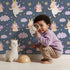 Lovely child standing in front of Whimsy Tots Unicorn Giggle Garden Wallpaper in Twilight Blue with unicorn, cloud, flower, heart in design.