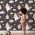 Lovely child standing in front of Whimsy Tots Unicorn Giggle Garden Wallpaper in Starry Night Black with unicorn, cloud, flower, heart in design.