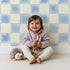 Lovely child standing in front of Whimsy Tots Gingham Watercolor Grid Wallpaper in Blueberry with checkerboard, square, watercolor in design.