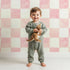Lovely child standing in front of Whimsy Tots Gingham Watercolor Grid Wallpaper in Strawberry with checkerboard, square, watercolor in design.
