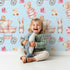 Lovely child standing in front of Whimsy Tots Tiny Tots Toy Parade Wallpaper in Sky with rabbit, bird, duck, bear, stroller, boot, toy, dress in design.
