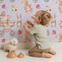 Lovely child standing in front of Whimsy Tots Tiny Tots Toy Parade Wallpaper in Lilac Lavender with rabbit, bird, duck, bear, stroller, boot, toy, dress in design.