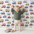 Lovely child standing in front of Whimsy Tots Tiny Wheels Parade Wallpaper in Soft Pink with car, truck, bus, firetruck, ambulance in design.