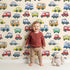Lovely child standing in front of Whimsy Tots Tiny Wheels Parade Wallpaper in Cream with car, truck, bus, firetruck, ambulance in design.