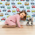 Lovely child standing in front of Whimsy Tots Tiny Wheels Parade Wallpaper in Sky Blue with car, truck, bus, firetruck, ambulance in design.