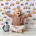 Lovely child standing in front of Whimsy Tots Bumper Buddies Rally Wallpaper in Soft Pink with car, truck, bus, foodtruck, taxi in design.
