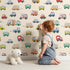 Lovely child standing in front of Whimsy Tots Bumper Buddies Rally Wallpaper in Cream with car, truck, bus, foodtruck, taxi in design.