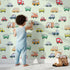 Lovely child standing in front of Whimsy Tots Bumper Buddies Rally Wallpaper in Mint Green with car, truck, bus, foodtruck, taxi in design.