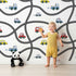 Lovely child standing in front of Whimsy Tots Bumper Buddies Road Maze Wallpaper in White with car, road, truck, taxi in design.