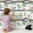 Lovely child standing in front of Whimsy Tots Bumper Buddies Town Tour Wallpaper in White with car, house, road, tree, stoplight in design.
