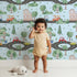 Lovely child standing in front of Whimsy Tots Bumper Buddies Town Tour Wallpaper in Soft Blue with car, house, road, tree, stoplight in design.