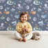 Lovely child standing in front of Whimsy Tots Undersea Slumber Voyage Wallpaper in Deep Blue with whale, submarine, octopus, boat, fish in design.