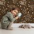 Lovely child standing in front of Whimsy Tots Playful Cub Spots Wallpaper in Brown with leopard, spot, pattern in design.
