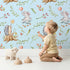 Lovely child standing in front of Whimsy Tots Starlight Forest Friends Wallpaper in Sky Blue with rabbit, hedgehog, star, flower, leaf in design.