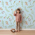 Lovely child standing in front of Whimsy Tots Blossom Meadow Breeze Wallpaper in Sky Blue with flower, leaf, vine, berry in design.