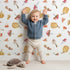 Lovely child standing in front of Whimsy Tots Autumn Forage Friends Wallpaper in White with mushroom, acorn, cherry, pumpkin, heart in design.