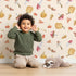 Lovely child standing in front of Whimsy Tots Autumn Forage Friends Wallpaper in Cream with mushroom, acorn, cherry, pumpkin, heart in design.