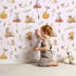 Lovely child standing in front of Whimsy Tots Wishful Woodland Friends Wallpaper in Blush Pink with hedgehog, owl, mouse, snail, mushroom, leaf, pumpkin, star in design.