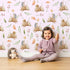Lovely child standing in front of Whimsy Tots Forest Stump Snuggle Wallpaper in Blush Pink with hedgehog, snail, tree stump, star, oak leaf, berry, leaf, mushroom in design.