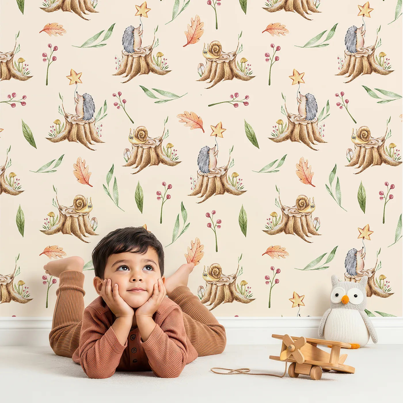 Lovely child standing in front of Whimsy Tots Forest Stump Snuggle Wallpaper in Cream with hedgehog, snail, tree stump, star, oak leaf, berry, leaf, mushroom in design.