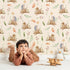 Lovely child standing in front of Whimsy Tots Forest Stump Snuggle Wallpaper in Cream with hedgehog, snail, tree stump, star, oak leaf, berry, leaf, mushroom in design.
