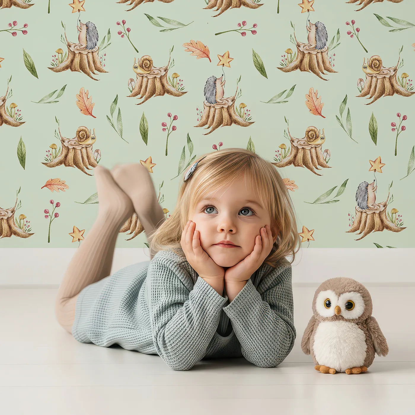 Lovely child standing in front of Whimsy Tots Forest Stump Snuggle Wallpaper in Green with hedgehog, snail, tree stump, star, oak leaf, berry, leaf, mushroom in design.