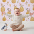 Lovely child standing in front of Whimsy Tots Pumpkin Patch Whimsy Wallpaper in Blush Pink with pumpkin, bird, heart, leaf in design.