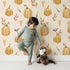 Lovely child standing in front of Whimsy Tots Pumpkin Patch Whimsy Wallpaper in Cream with pumpkin, bird, heart, leaf in design.
