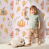 Lovely child standing in front of Whimsy Tots Pumpkin Heart Harvest Wallpaper in Blush Pink with pumpkin, mushroom, leaf, heart, berry in design.