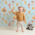 Lovely child standing in front of Whimsy Tots Pumpkin Heart Harvest Wallpaper in Sky Blue with pumpkin, mushroom, leaf, heart, berry in design.