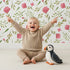 Lovely child standing in front of Whimsy Tots Blushing Petal Tangle Wallpaper in White with flower, leaf, petal, vine in design.
