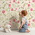 Lovely child standing in front of Whimsy Tots Blushing Petal Tangle Wallpaper in Cream with flower, leaf, petal, vine in design.