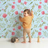 Lovely child standing in front of Whimsy Tots Blushing Petal Tangle Wallpaper in Sky with flower, leaf, petal, vine in design.