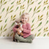 Lovely child standing in front of Whimsy Tots Wandering Willow Sprigs Wallpaper in Cream with leaf, sprig, branch in design.